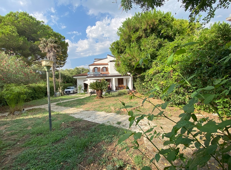 A spacious garden surrounds the villa, featuring well-maintained greenery and natural landscaping. A stone pathway leads to the entrance of the two-story house, which is framed by trees and shrubs. A car is parked near the front, adding to the inviting exterior.