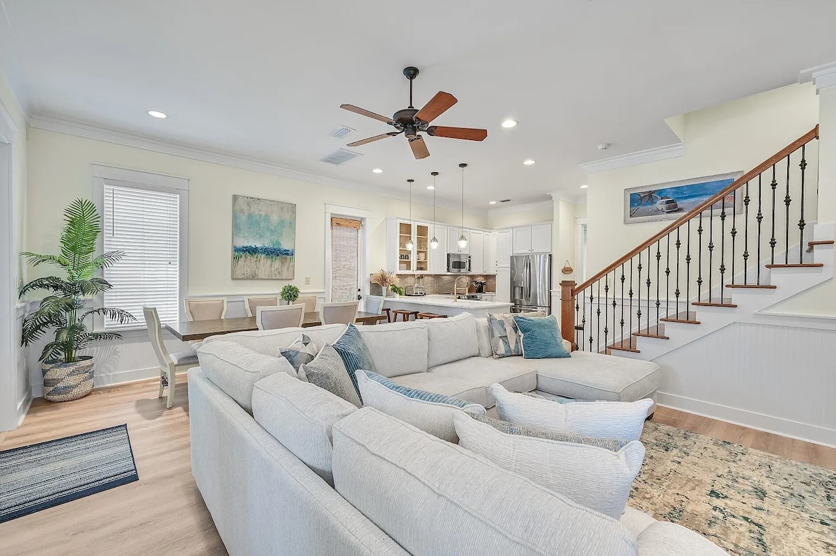 An inviting living area features a large sectional sofa with neutral upholstery, complemented by decorative pillows. A ceiling fan hangs above, while a dining table is visible in the background. Stairs lead to the upper level, framed by a decorative railing, and natural light fills the space.