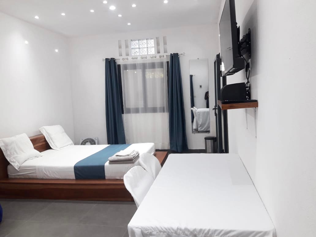 A well-lit room features a spacious bed with white linens and a blue accent blanket. A clean table is situated nearby, complemented by a modern television mounted on the wall. Large windows are dressed with dark curtains, allowing natural light to fill the space.