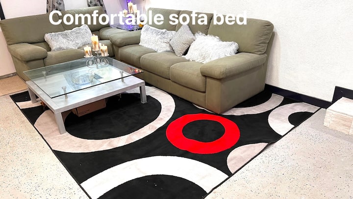 „Отпуснете се в тази уютна всекидневна с плюшен матрак в маслиненозелен цвят, пухкави възглавници, модерна стъклена масичка за кафе и смел дизайнерски килим с червени, черни и бели шарки. Перфектно място за отдих или удобен сън.“