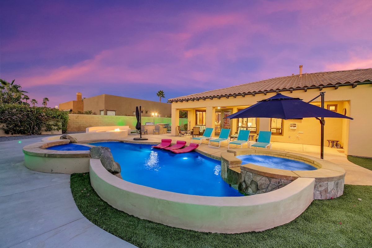 An inviting outdoor space highlights a heated saltwater pool and spa, surrounded by multiple lounge chairs and colorful pool floats. A shaded area is provided by an umbrella, while the sunset casts a soft glow over the scene, enhancing the tranquil ambiance.