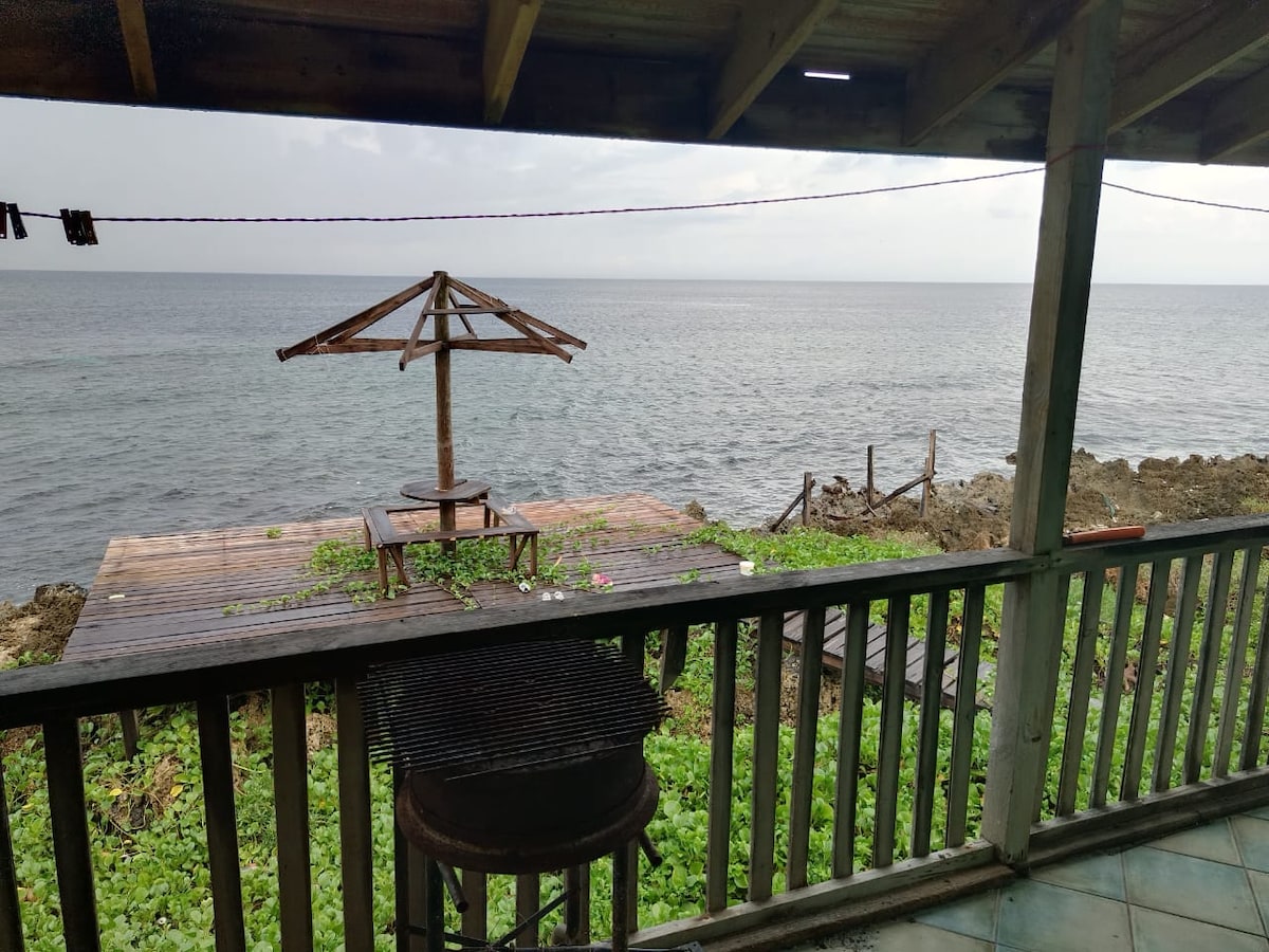 A wooden deck extends over rocky terrain, overlooking calm waters. A weathered gazebo stands in one corner, while a grill is positioned prominently on the balcony. Lush green vegetation surrounds the space, enhancing the natural coastal setting.