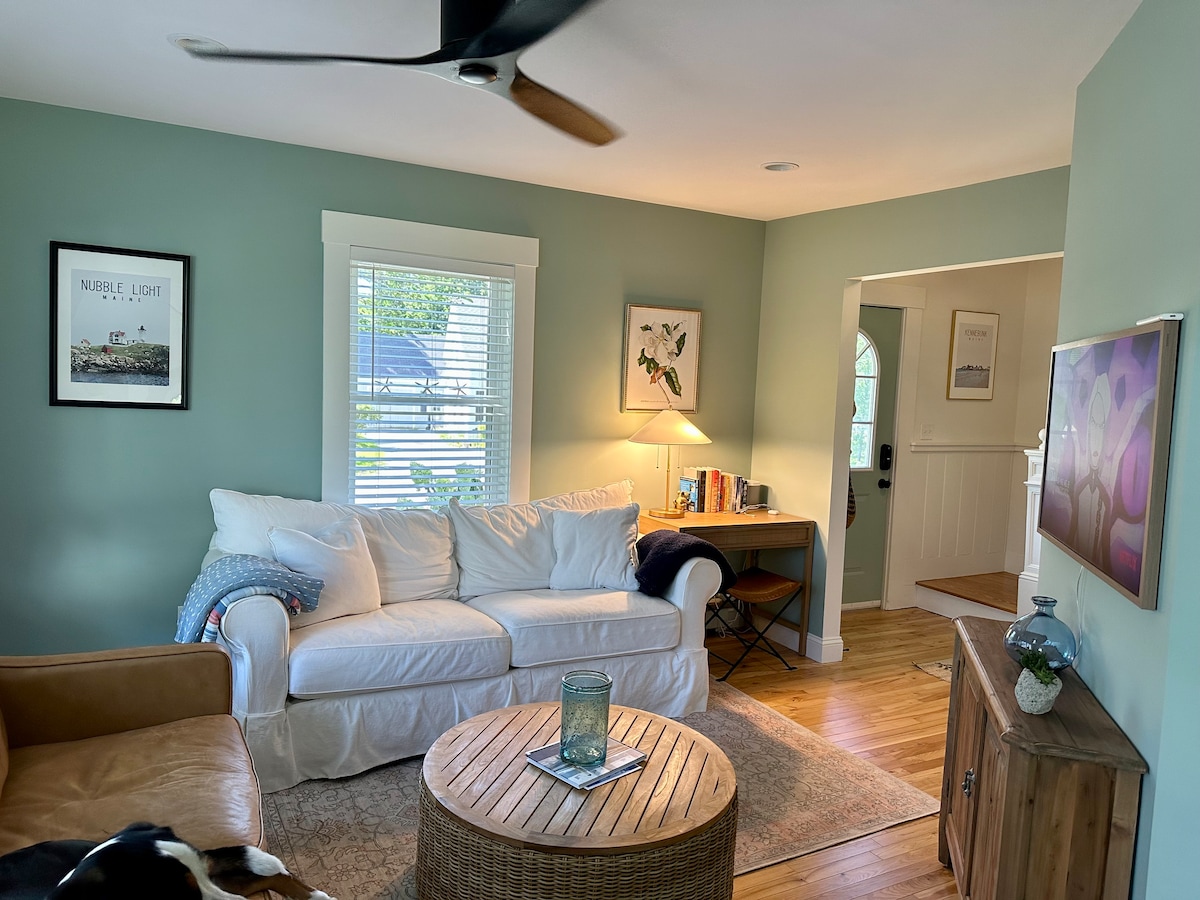 A cozy living room features a light-colored sectional sofa with a textured throw and a round woven coffee table. A fan hangs from the ceiling. Natural light filters through the window, illuminating the space. A desk with books and artwork is positioned near the entrance.
