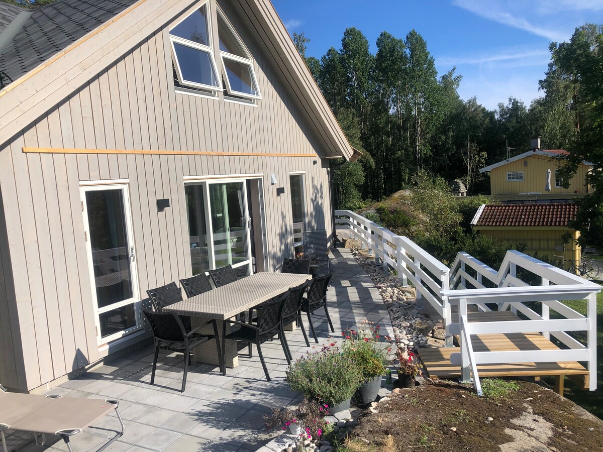 An outdoor patio features a table with chairs, surrounded by greenery and flowers. The wooden structure showcases large windows, and a pathway leads to a side deck with a railing. The setting is illuminated by natural light under a clear blue sky.