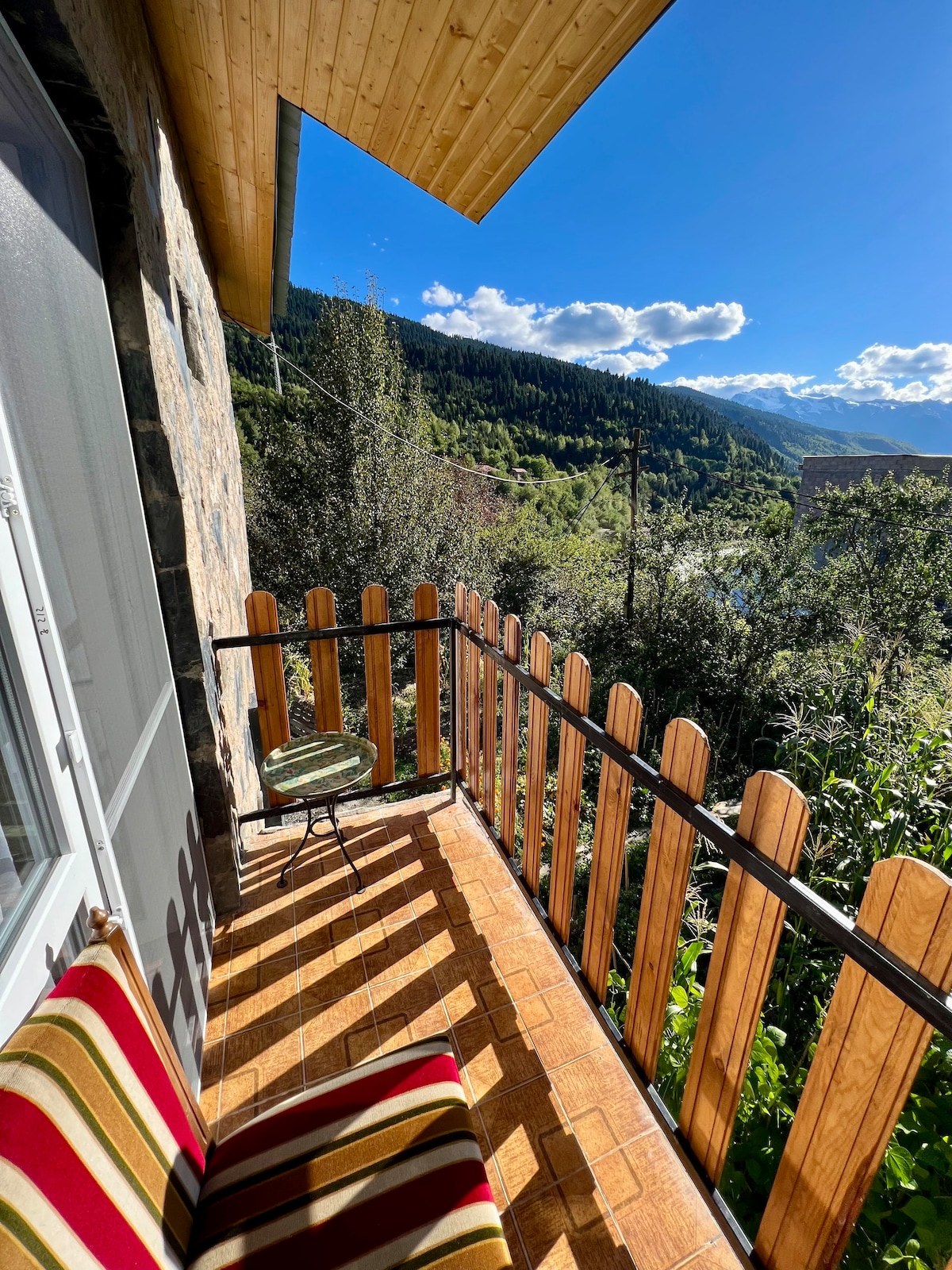 A small balcony is framed by wooden fencing, overlooking lush greenery and distant mountains under a clear blue sky. A patterned striped cushion adorns a bench, while a round table stands nearby, enhancing the outdoor space's comfort and connection to nature.