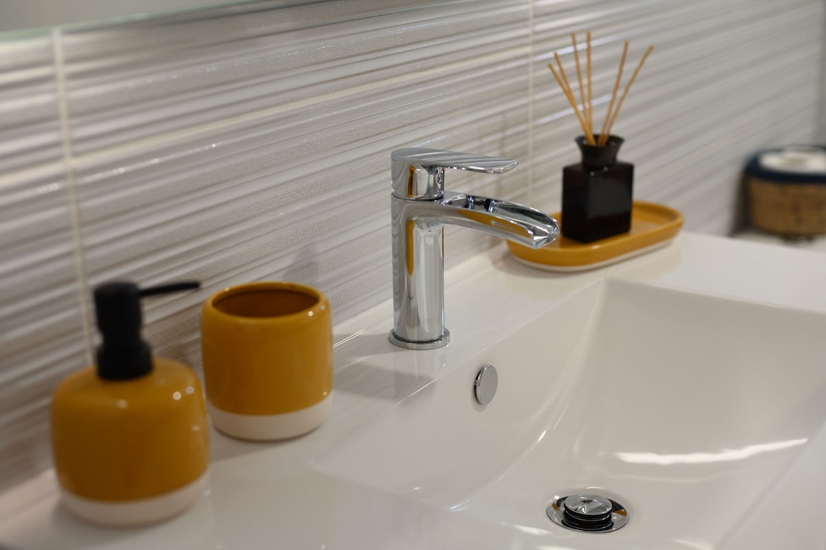 A modern bathroom sink is presented, featuring a sleek chrome faucet. To the left, two yellow containers add a pop of color, while a small black vase with reed diffusers is positioned on a rectangular tray at the back.