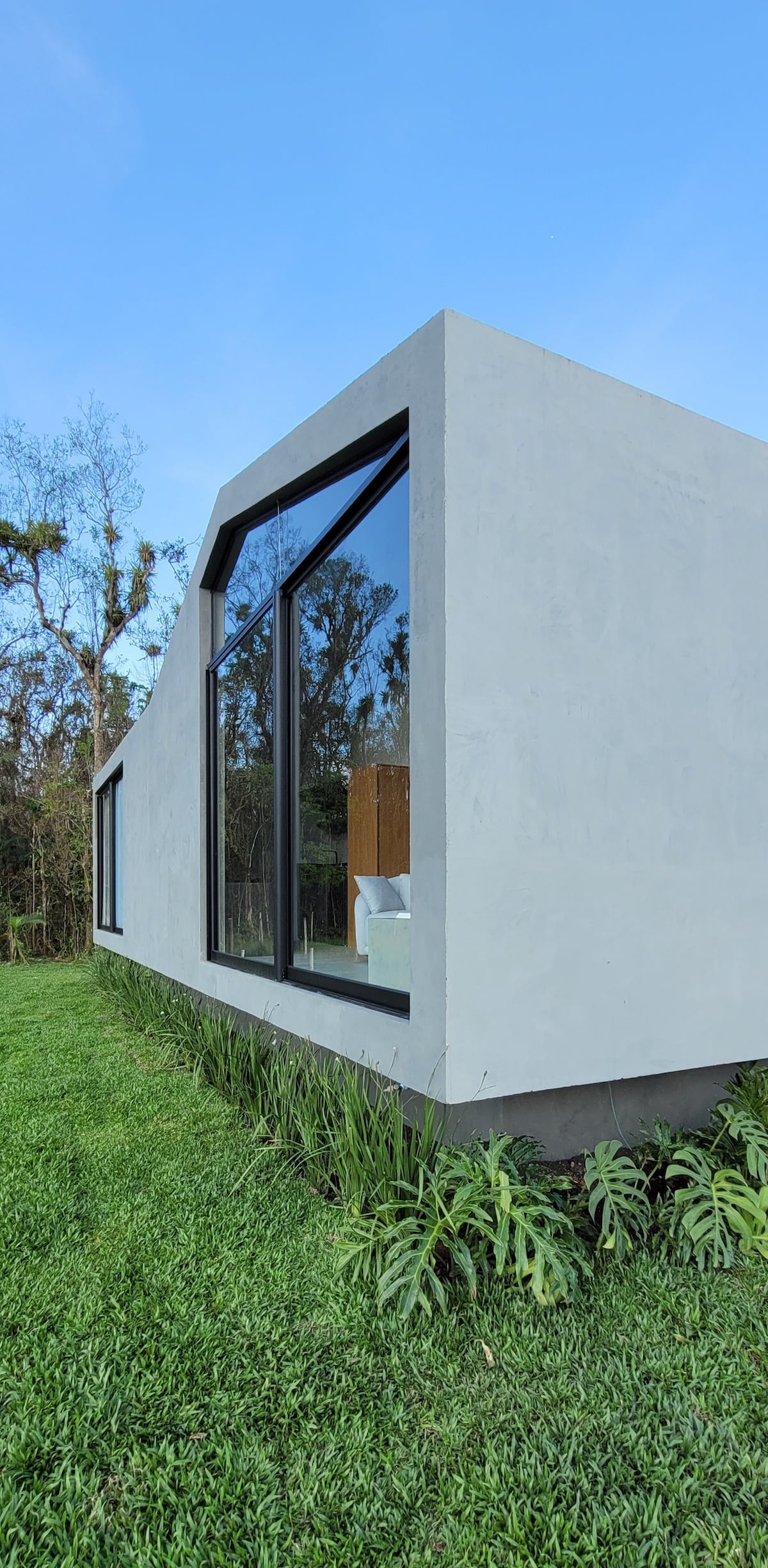 A modern structure is shown from an exterior angle, featuring large windows that offer natural light. The building is surrounded by lush greenery, with neatly trimmed grass and a border of plants highlighting the design. The setting appears peaceful and integrated with nature.