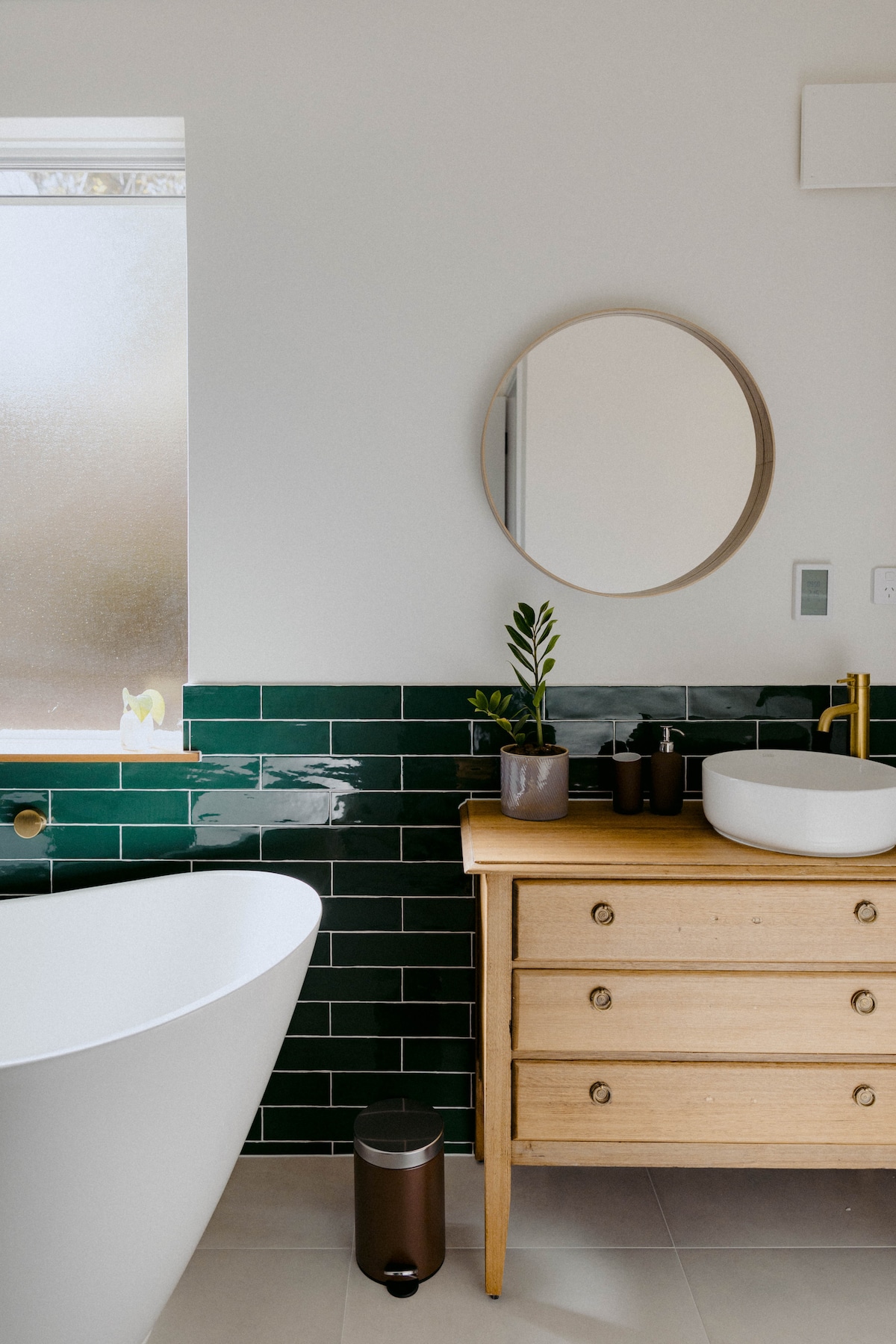 A modern bathroom features a freestanding soaking tub beside a wooden vanity with a round mirror. Green and white tiled walls are complemented by natural light filtering through a frosted window. A small potted plant adds a touch of greenery to the space.