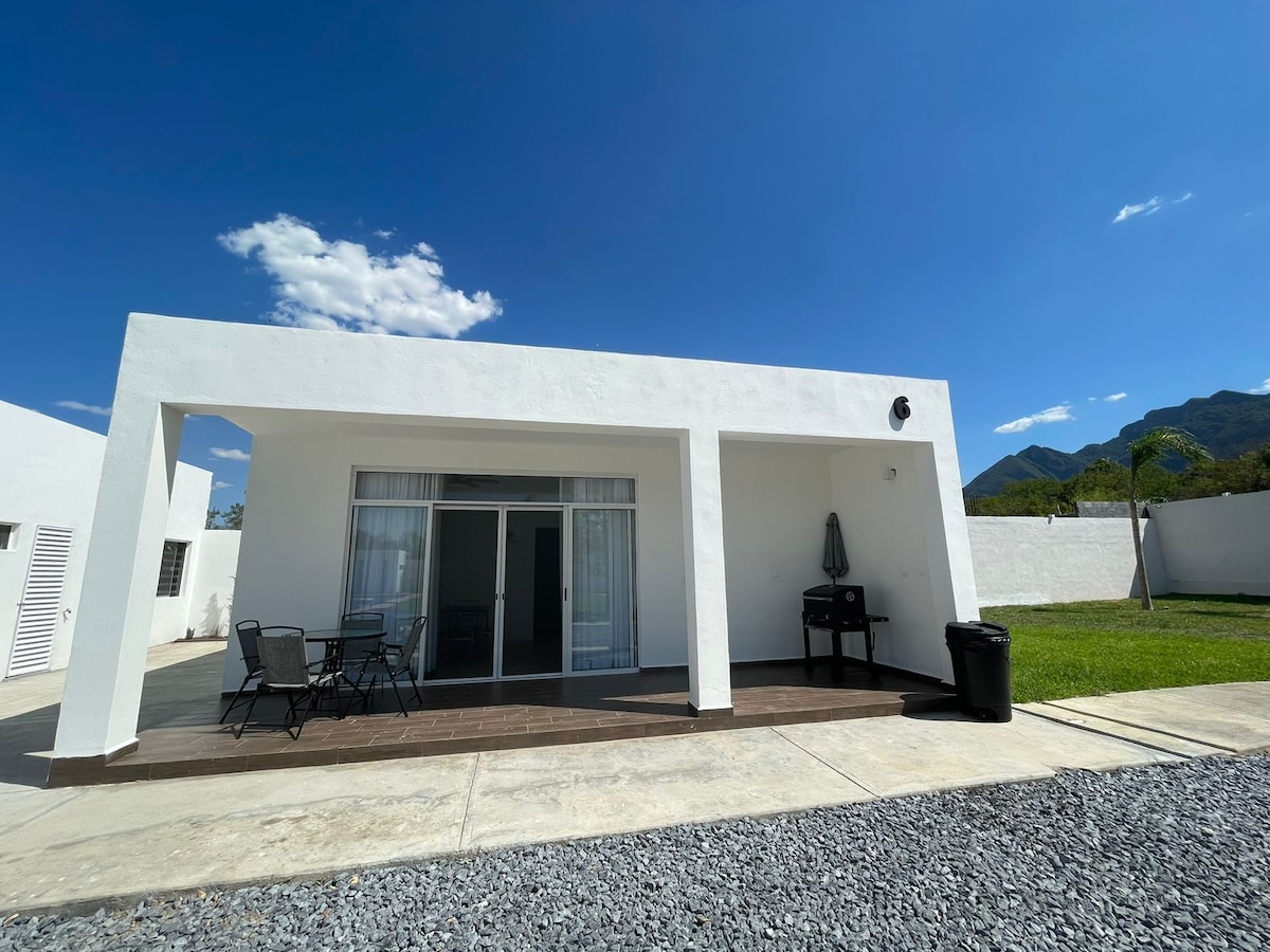 The exterior of the villa is presented with a modern design featuring a large covered patio. A dining table with chairs occupies one side, while a grill is placed nearby. The surrounding landscape includes a well-maintained lawn and mountains visible in the background.
