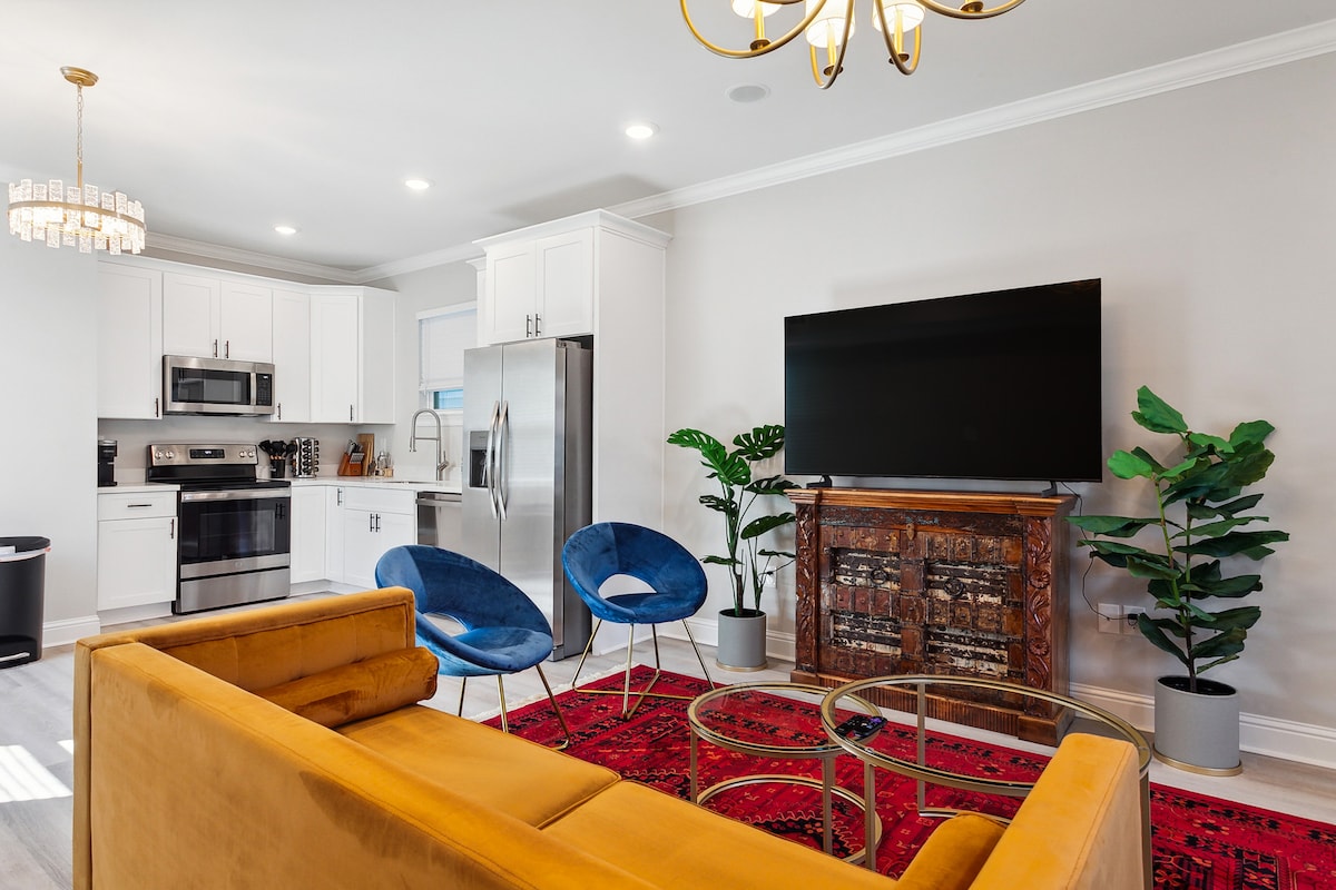 A modern living area features a yellow sofa facing a large, mounted television. A patterned area rug anchors the space, complemented by two blue accent chairs. A dining table is situated nearby, and potted plants add a touch of greenery to the room.