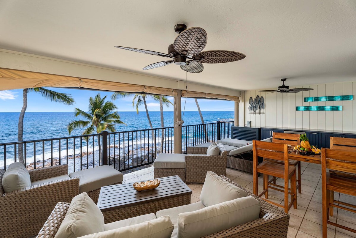 A spacious covered lanai features comfortable seating arrangements and a dining area overlooking the ocean. The view includes palm trees swaying gently in the breeze, while the sound of waves enhances the inviting atmosphere. Natural light fills the area through expansive windows.