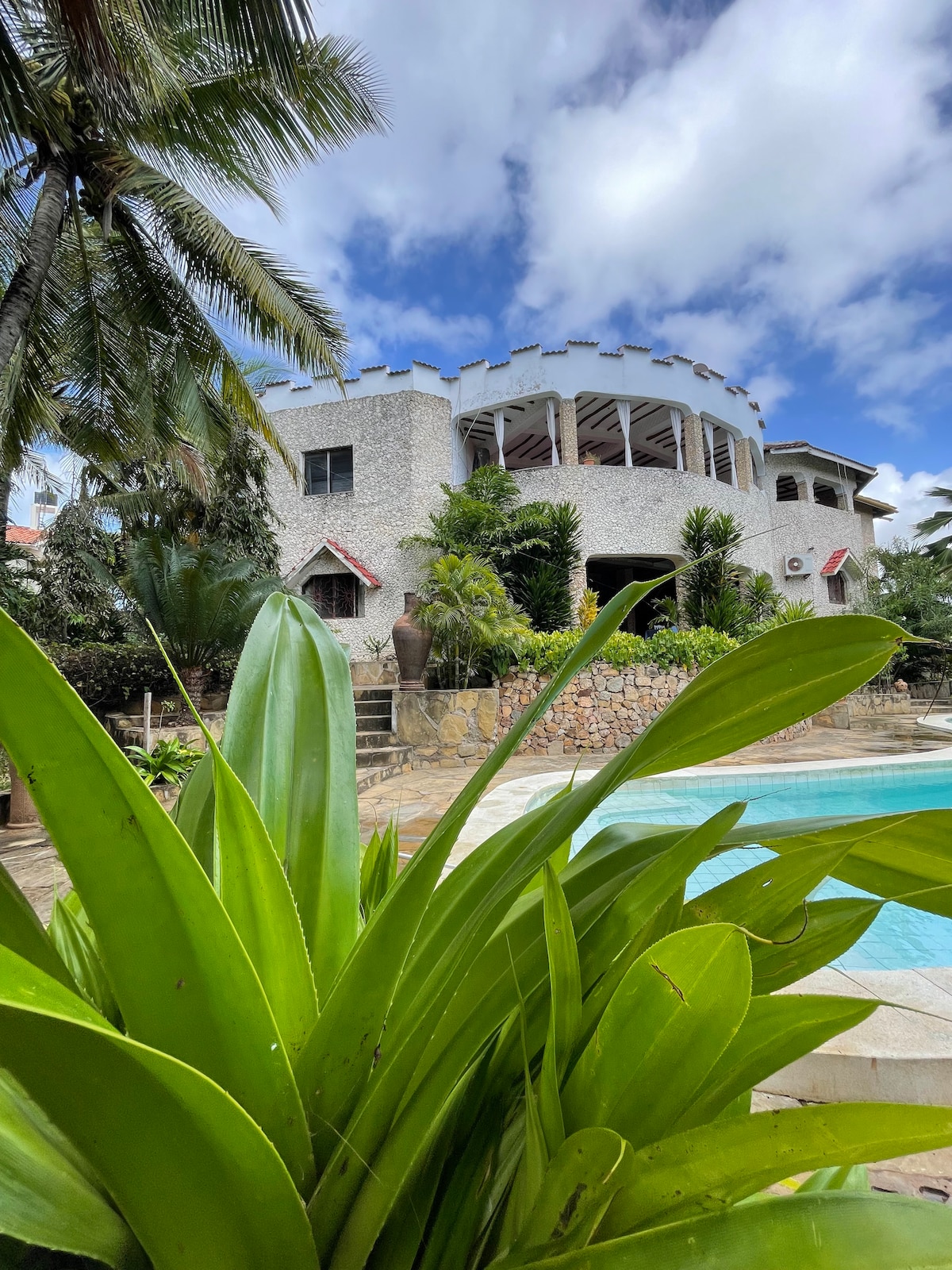 A spacious multi-level structure is framed by lush green foliage and palm trees, showcasing a unique architectural design. The exterior features stone walls and an array of windows. A large pool can be seen in the foreground, surrounded by a well-maintained garden.