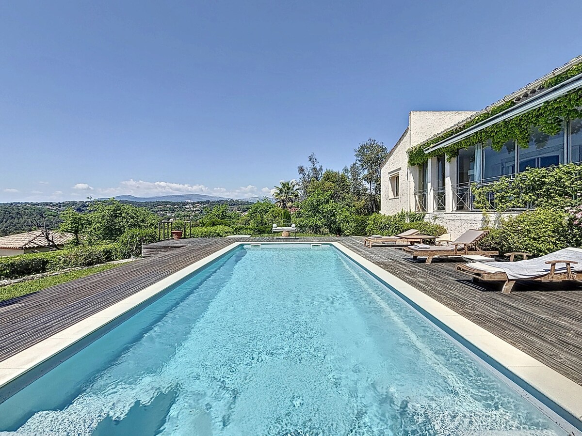 A spacious outdoor area features a clear blue swimming pool surrounded by wooden decking. Sun loungers are situated along the edge, and lush greenery frames the scene, enhancing the view of the hills in the distance under a bright sky.