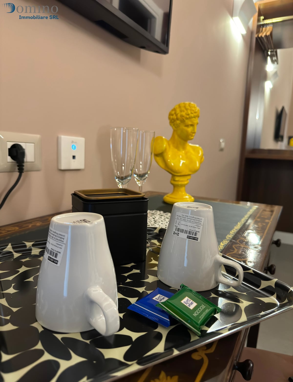A tray displays two white cups, a small black container, and green and blue tea packets. Behind, a pair of clear glasses rests beside a yellow decorative bust. A dark wooden surface with intricate patterns is visible in the background.