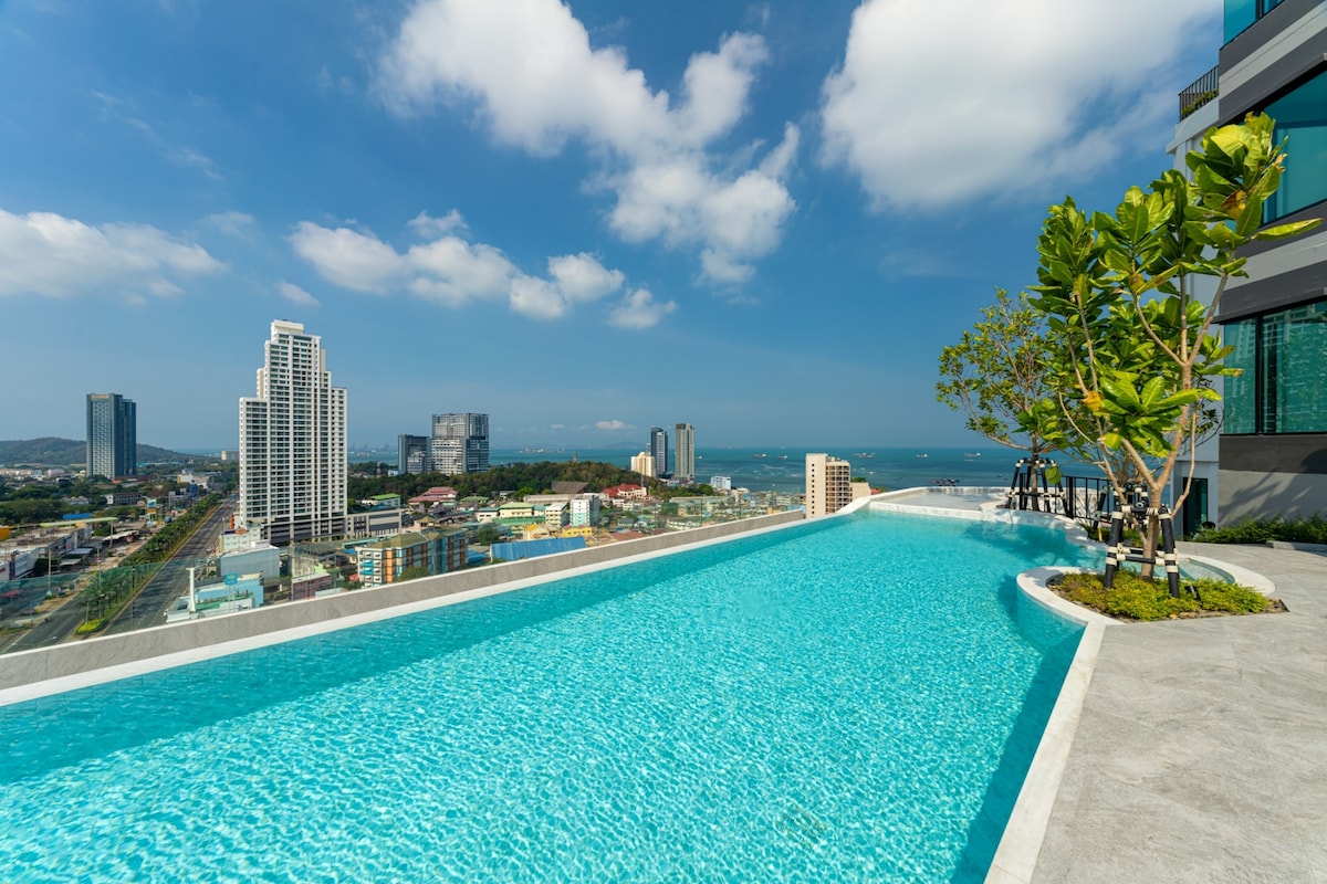 An infinity pool extends towards the horizon, revealing a panoramic view of the cityscape and ocean. Lush greenery is positioned along the edge, complementing the clear blue water under a bright sky dotted with clouds.