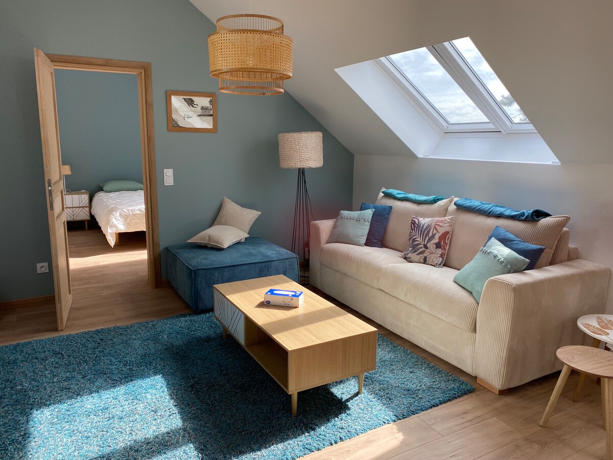 A cozy living area features a light-colored sofa adorned with assorted cushions, complemented by a wooden coffee table and an ottoman. The space is illuminated by natural light streaming through skylights and a nearby window, with a large area rug enhancing the comfort of the room.