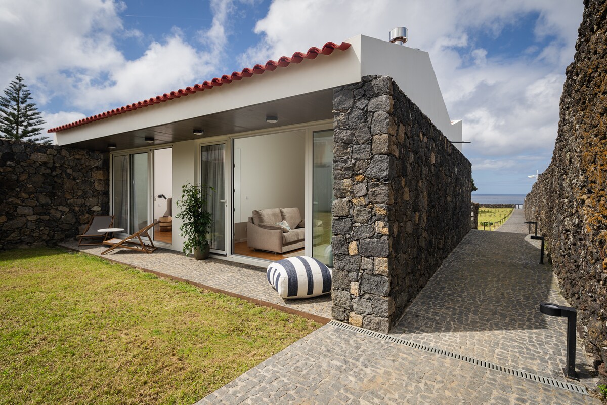A modern structure is framed by natural stone walls, featuring large glass doors that open to a grassy area. Comfortable seating, including a light-colored sofa and an outdoor lounging chair, is positioned nearby, inviting relaxation. The sky is partly cloudy, enhancing the serene atmosphere.