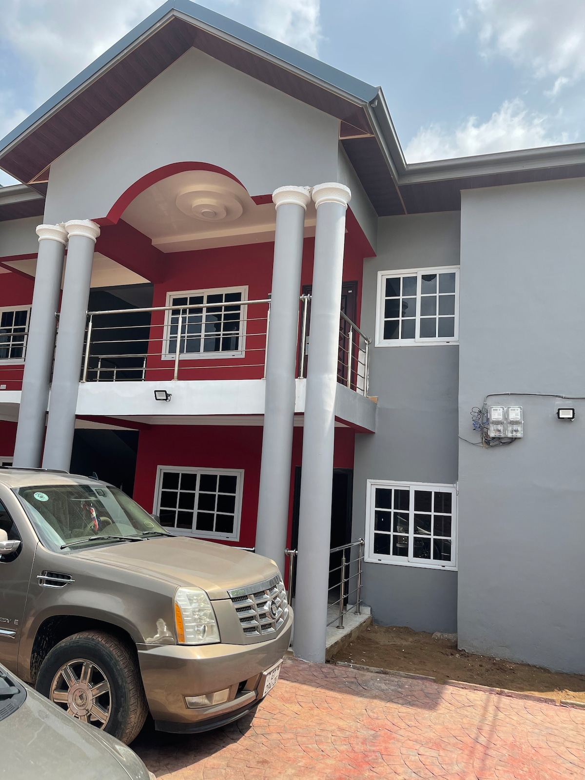 A modern two-story building features a combination of red and gray exteriors. Large windows allow natural light to enter, and tall columns support a covered entrance. A parked vehicle is visible in the foreground, and the surrounding area appears well-maintained.