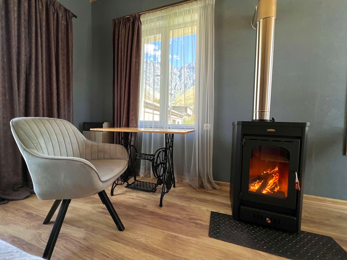 A cozy corner of the studio apartment features a woodburning fireplace, casting warmth. A stylish chair sits adjacent to a simple table, with natural light filtering through sheer curtains. The mountain views are visible through the large window, enhancing the serene atmosphere.