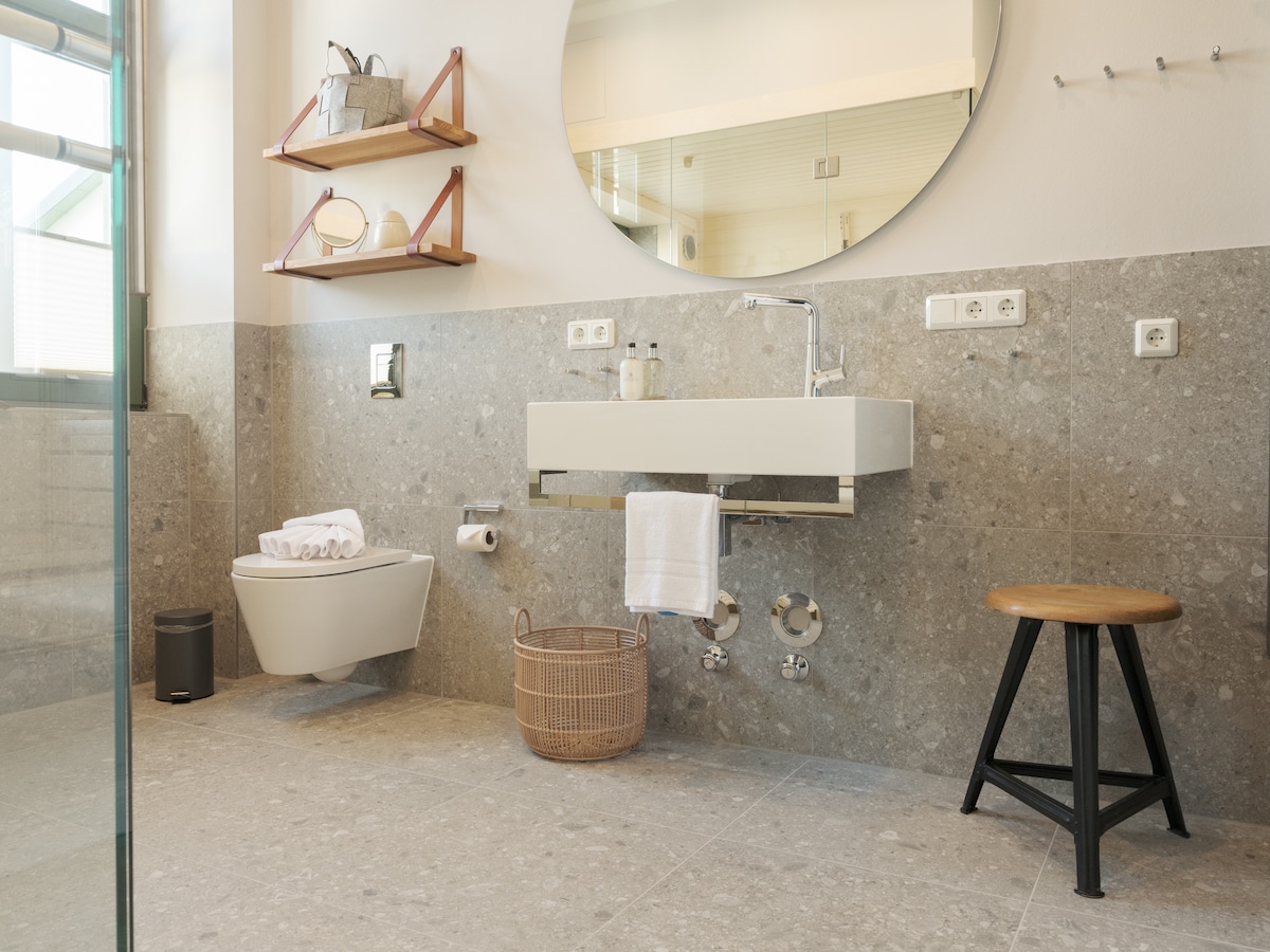 A modern bathroom features a wall-mounted sink with overhead storage shelves. Natural stone tiles cover the floor and lower walls. A round mirror reflects light, while a wooden stool offers additional seating. A woven basket and neatly arranged towels enhance the functional design.