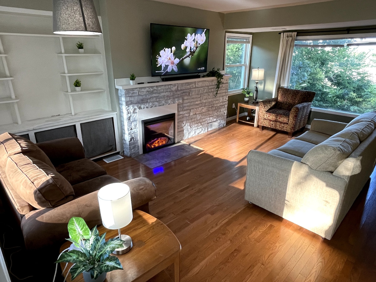 A comfortable living room is presented, featuring a cozy couch and an armchair. A fireplace adds warmth, while a large television is mounted above a stone mantel. Natural light streams through the window, highlighting the wooden flooring and decorative plants on the shelves.