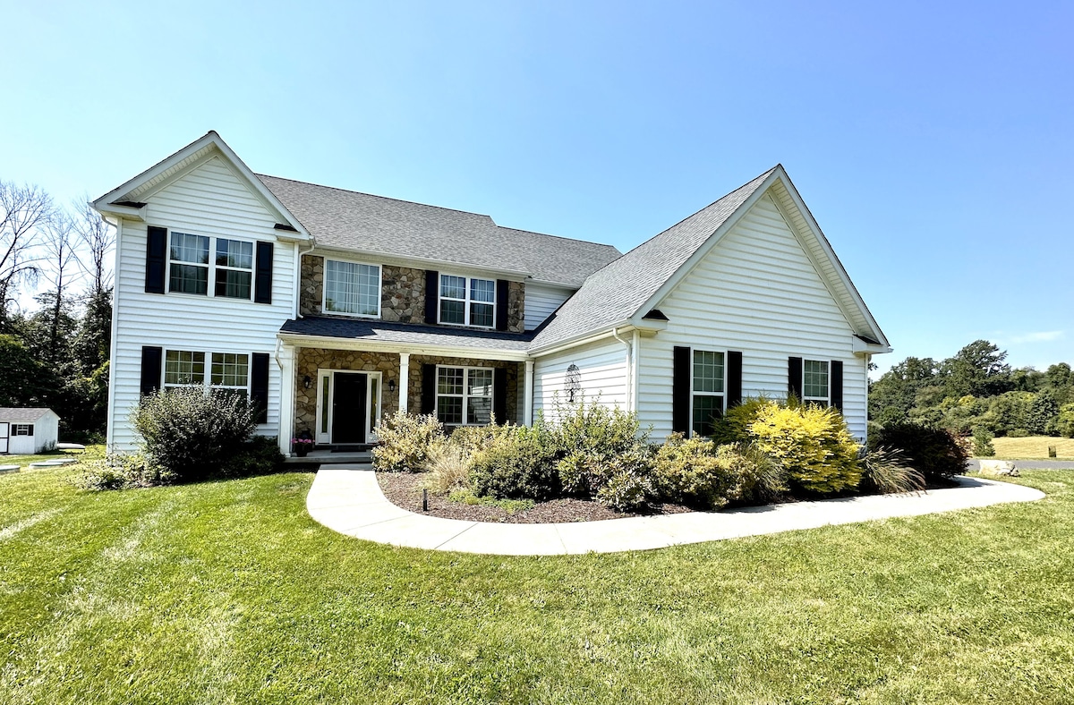 A spacious two-story home features a combination of white siding and stone accents. The well-maintained landscaping includes shrubs and flower beds surrounding a curved walkway leading to the front entrance, all set against a clear blue sky.