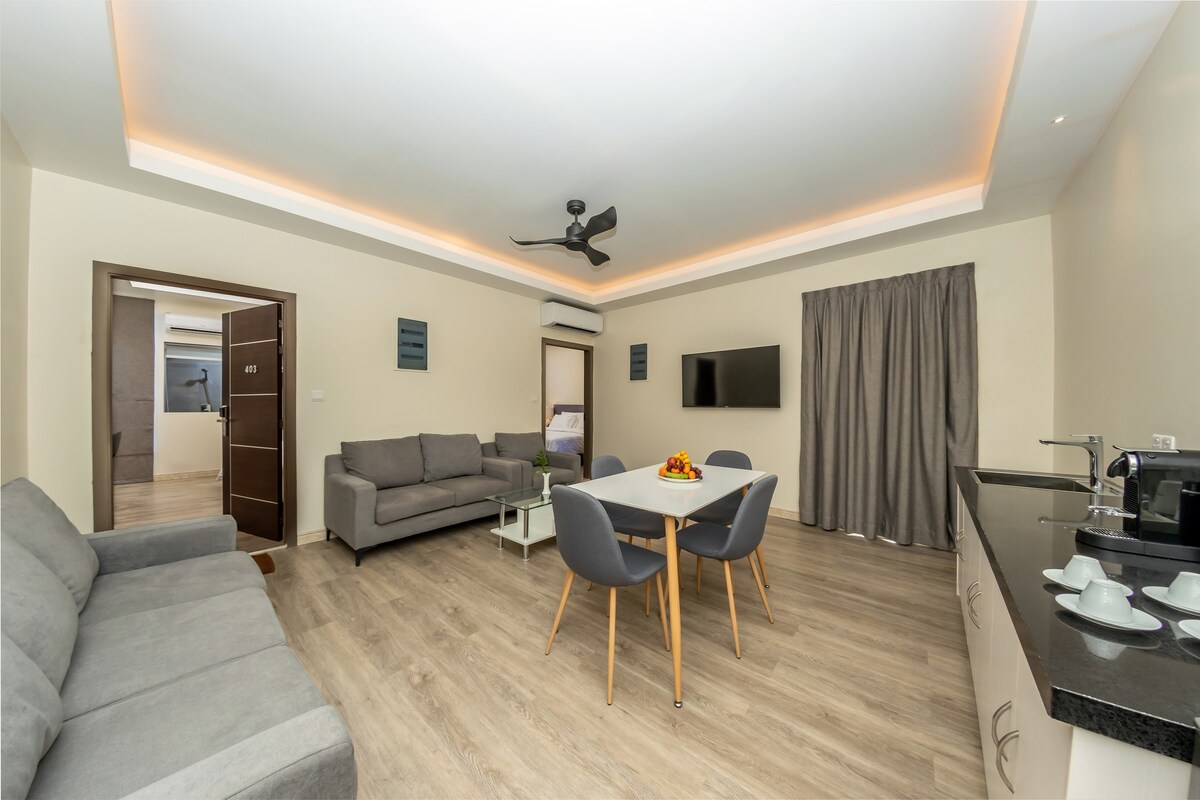 A spacious living area is featured, furnished with two grey sofas and a dining table set for four. Natural light fills the space through a window, and a ceiling fan enhances comfort. A kitchenette with modern appliances is present, offering a functional area for meal preparation.