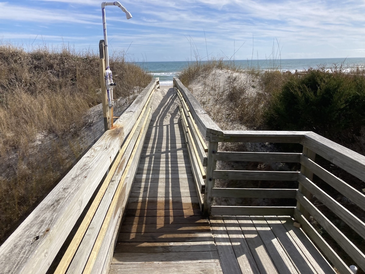 A wooden boardwalk leads from the home to the beach, surrounded by natural vegetation. A shower is positioned on the left side, and the ocean is visible in the distance beyond the sandy dunes, inviting beach access under a clear blue sky.