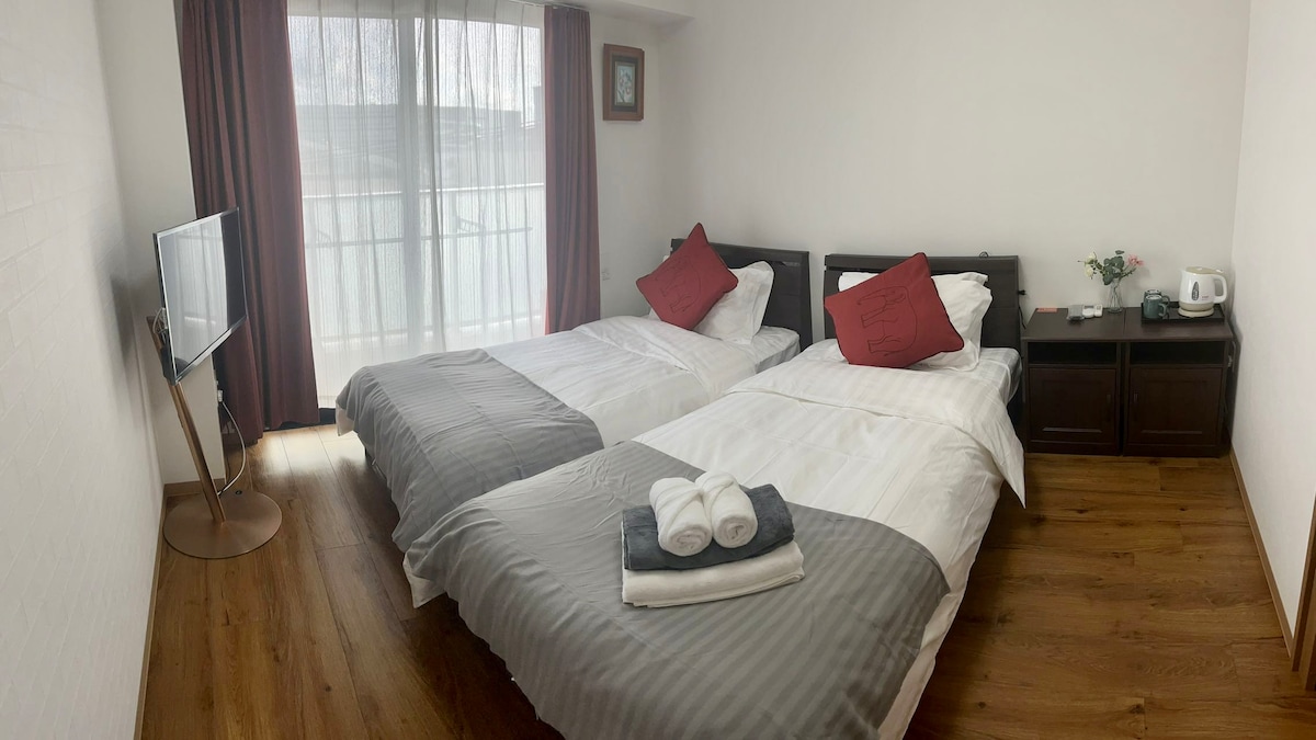 A well-appointed guest room features two spacious beds, each adorned with decorative pillows. A TV is positioned on a low stand, while a small table holds a kettle and a vase, adding subtle details. Natural light filters through sheer curtains, creating a bright atmosphere.