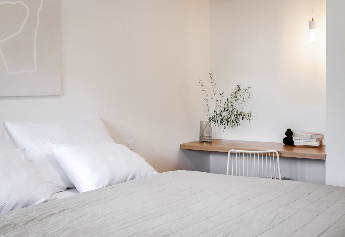 A modern sleeping area features a spacious bed with light-colored bedding and soft pillows. A simple wooden desk is positioned nearby, adorned with a small vase containing greenery. A warm light bulb hangs overhead, illuminating the serene space.