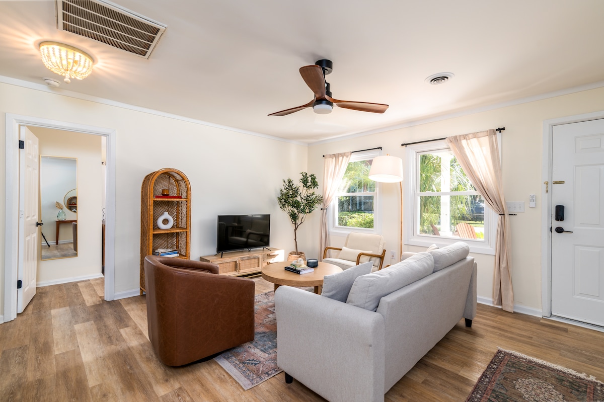 The living area features a comfortable sofa and a brown armchair arranged around a central coffee table. A TV stands on a media console, flanked by indoor plants. Natural light fills the room through two windows, complemented by soft curtains, enhancing the inviting atmosphere.