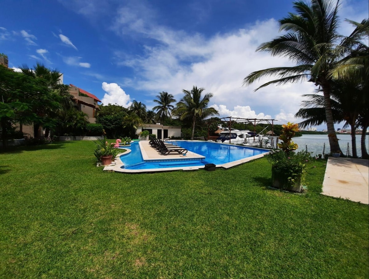 A well-maintained garden surrounds a large swimming pool, featuring several lounge chairs positioned for relaxation. Palm trees sway gently under a clear blue sky, while a private dock is visible along the lagoon's edge, creating a serene outdoor atmosphere.