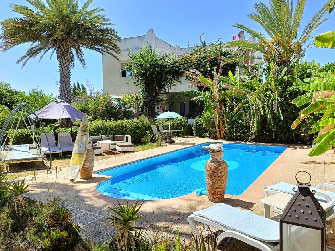 A well-kept garden surrounds a tranquil swimming pool, with palm trees and lush greenery providing shade. Comfortable lounge chairs are positioned nearby, alongside a hammock and a parasol. The pool's clear blue water reflects the sunlight, inviting relaxation in a serene outdoor space.