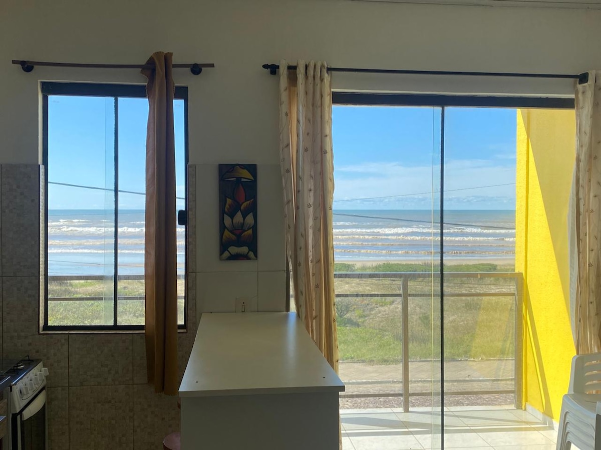 Large windows frame a view of the ocean, allowing natural light to fill the interior. A bright yellow wall contrasts with light-colored cabinetry, while a white countertop provides a functional space. Sheer curtains add texture, softening the view of the shoreline.