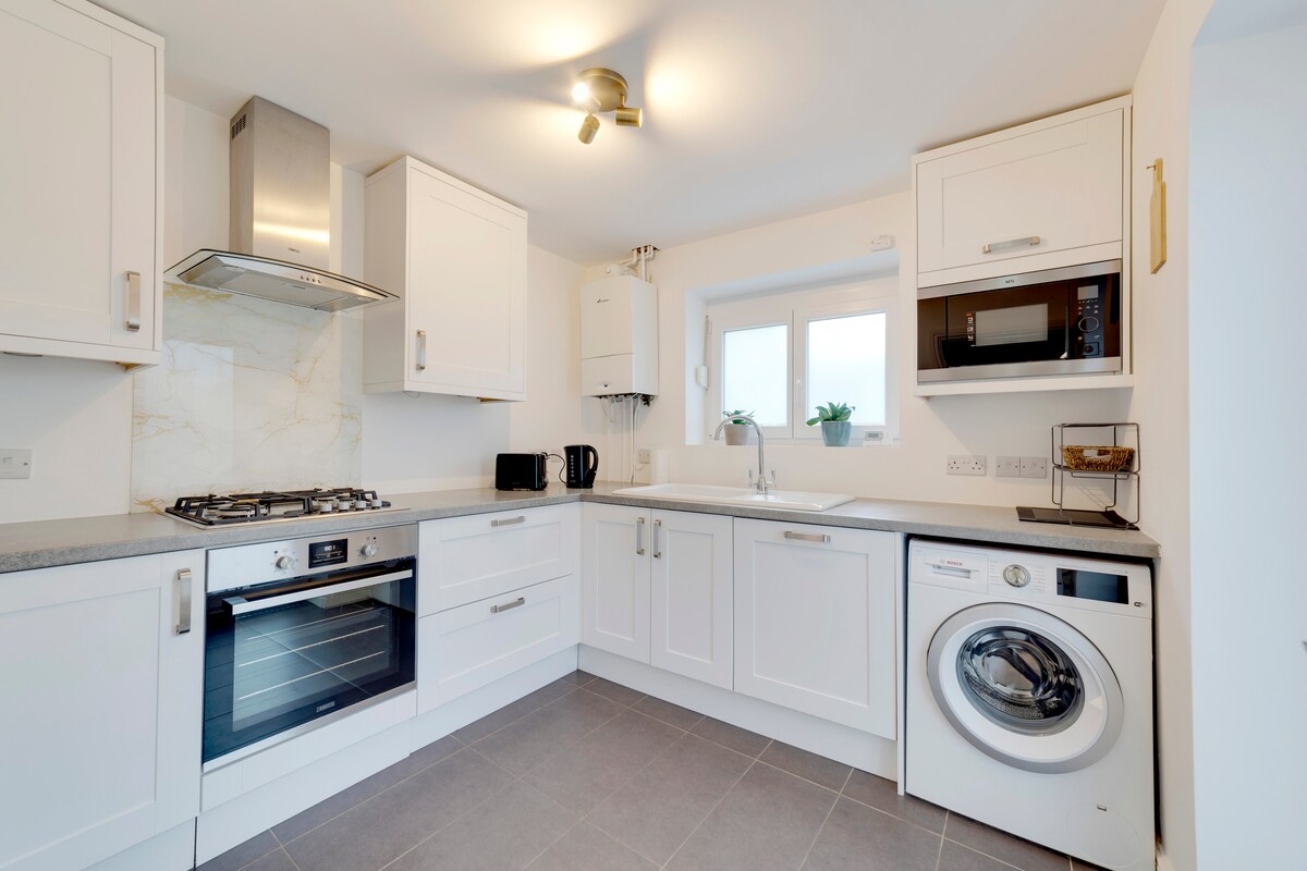 The modern kitchen features white cabinetry and granite countertops. Appliances include a gas oven, microwave, kettle, and a washer/dryer. Natural light enters through a window above the sink, complemented by potted plants. A clean, minimalistic layout enhances functionality for home dining.