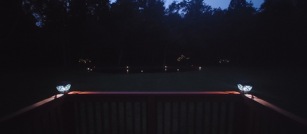 The deck offers a serene view of the expansive yard illuminated by soft outdoor lighting. Decorative lanterns outline the landscape, while the dark silhouette of trees stands in the background. A small pond is faintly visible, enhancing the peaceful nighttime atmosphere.