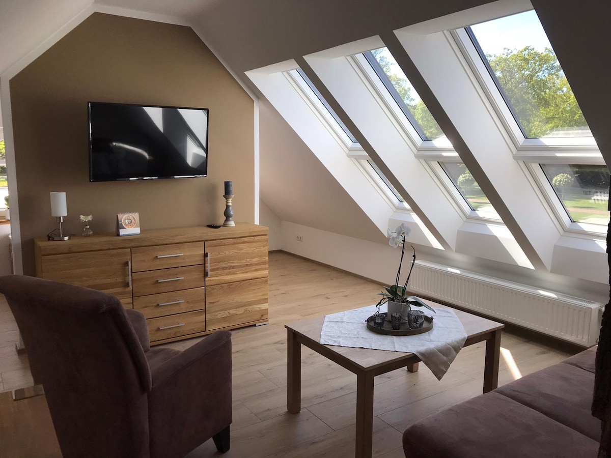 A bright living area is illuminated by large skylights, offering views of the greenery outside. A wooden sideboard sits against the wall, complemented by a small table with a decorative plant. Comfortable seating is arranged around a central table, creating an inviting space.