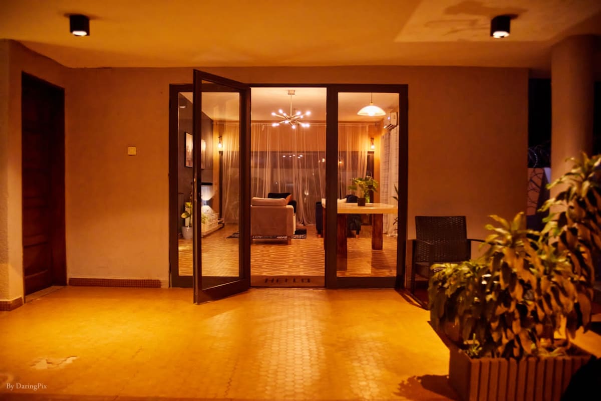 An entryway features double glass doors leading to a bright living area. Inside, a cozy seating arrangement is visible, illuminated by soft lighting. Potted plants add greenery to the exterior, while warm tile flooring contributes to the inviting ambiance.