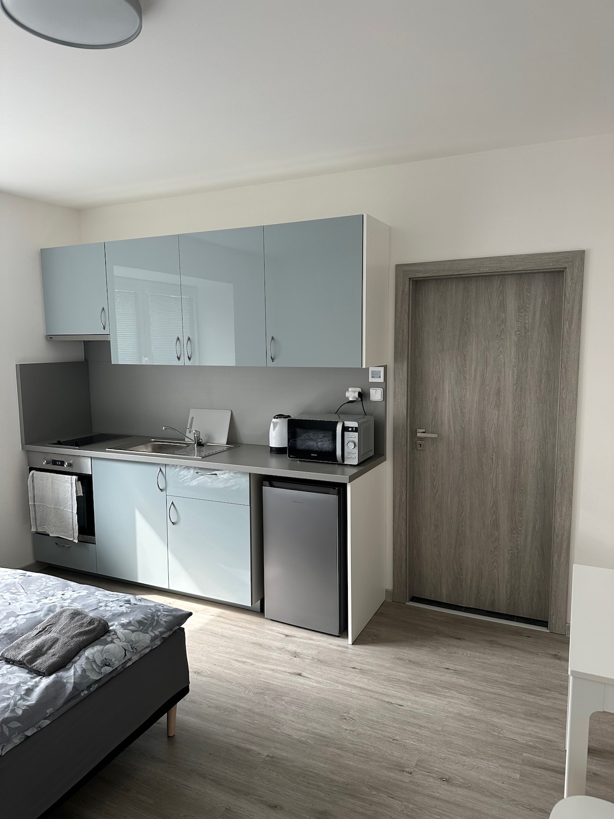 A compact kitchenette features blue cabinetry and a gray countertop. Appliances such as a microwave and refrigerator are visible. A door to another room is present in the background, while natural light enters through a nearby window.