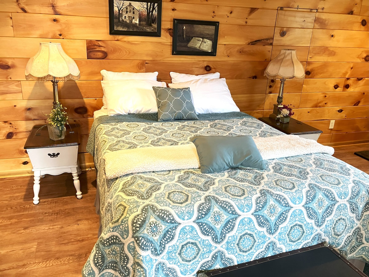 A cozy sleeping area features a spacious bed dressed in a patterned blue quilt. Flanking the bed are two lamps on nightstands, with decorative plants adding a personal touch. Warm wooden walls create a welcoming environment.