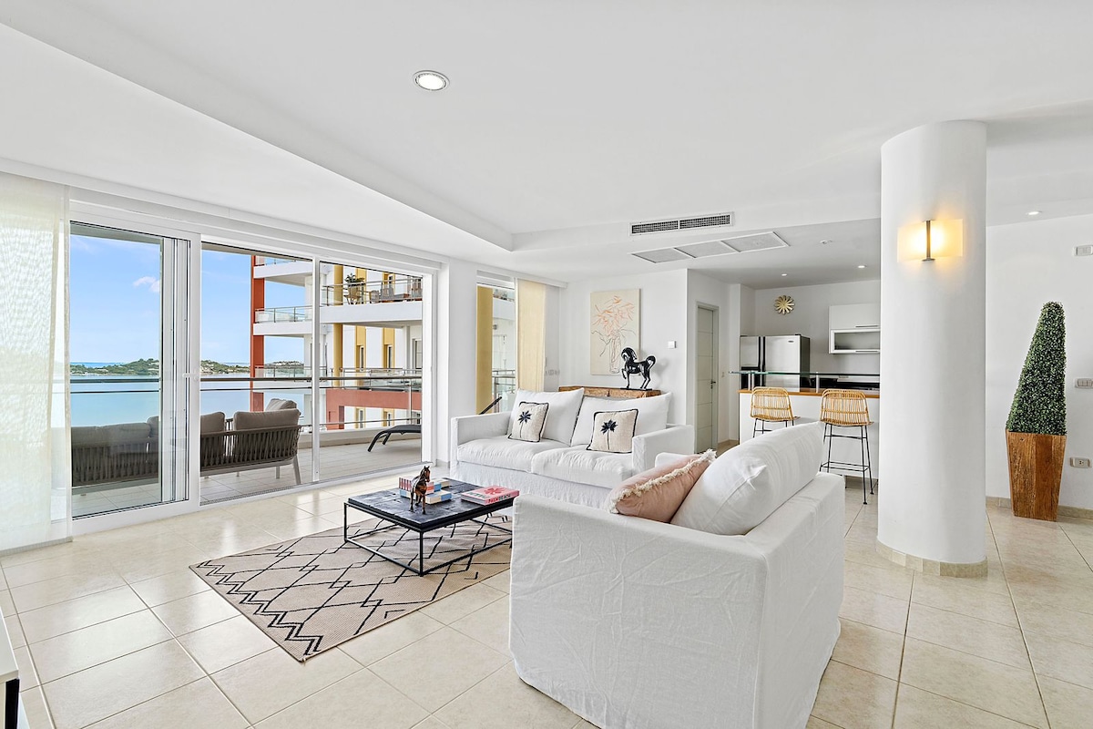An open living area features light-colored sofas with decorative cushions and a low coffee table, surrounded by large glass doors that offer views of the water. A modern kitchen with stainless steel appliances is visible in the background, alongside a dining area with stylish bar stools.