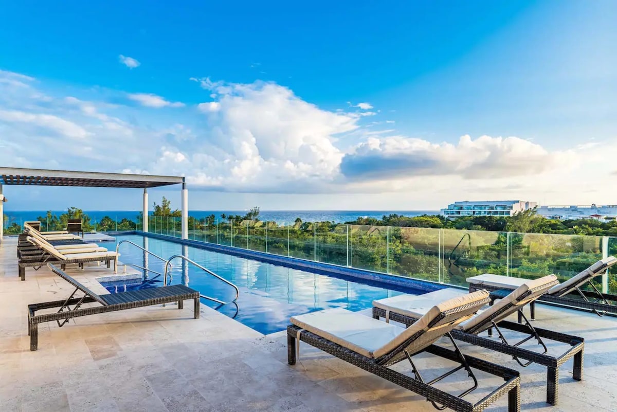 A rooftop pool area is presented with lounge chairs arranged to enjoy panoramic views of the ocean and surrounding landscape. The tranquil water reflects the sky, and a shaded area provides comfort. Lush greenery and distant buildings can be seen in the background.
