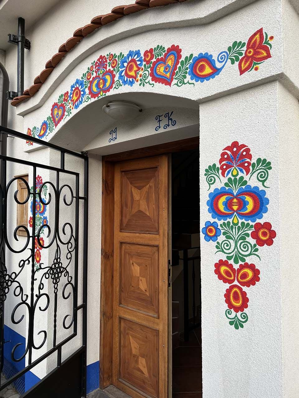 The entrance features a wooden door adorned with intricate carvings and vibrant floral patterns. Colorful decorative motifs enhance the surrounding wall, adding a touch of local artistry. A wrought iron gate offers security while complementing the welcoming appearance of the entrance.