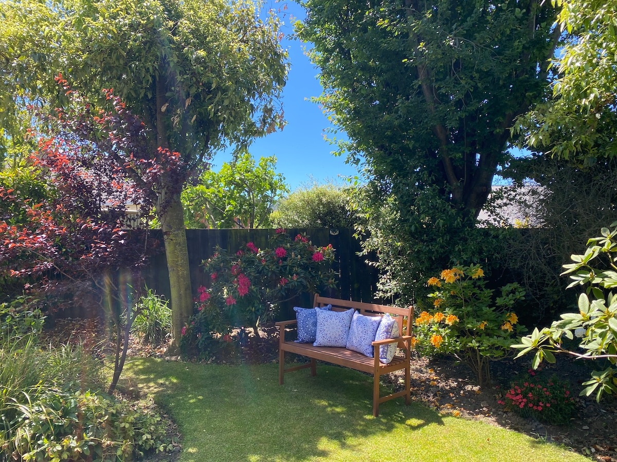 A serene garden setting is visible, featuring a wooden bench adorned with cushions, surrounded by lush greenery and colorful flowering plants. Tall trees offer shade, creating a peaceful retreat that invites relaxation.