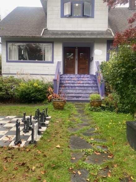 The exterior of a charming heritage house features a set of wooden front doors, approached by a short set of purple-painted stairs. Lush greenery frames the entrance, complemented by decorative planters. A large outdoor chess set set on a grassy area adds a unique touch.