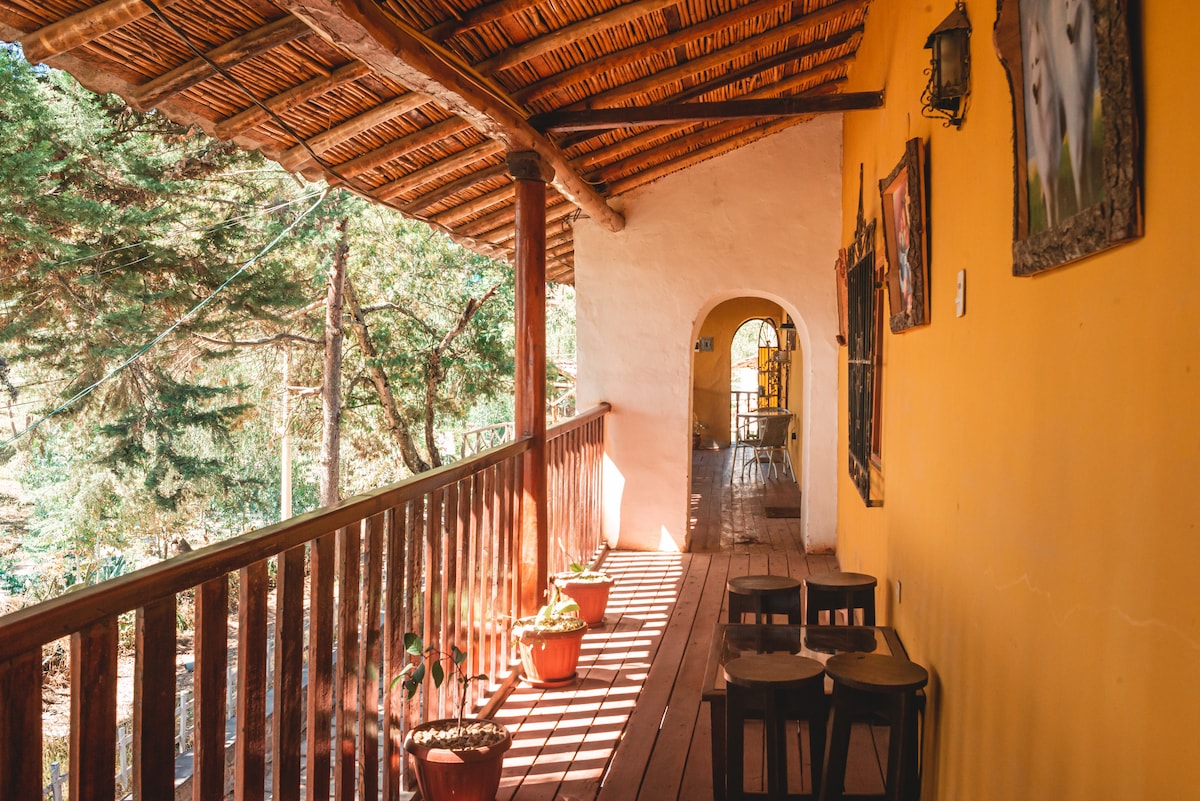 A covered balcony features a warm yellow wall and wooden flooring, with potted plants placed alongside. Small wooden tables and chairs are arranged for seating, surrounded by a serene forest view visible through the balustrade and archway leading to the interior.