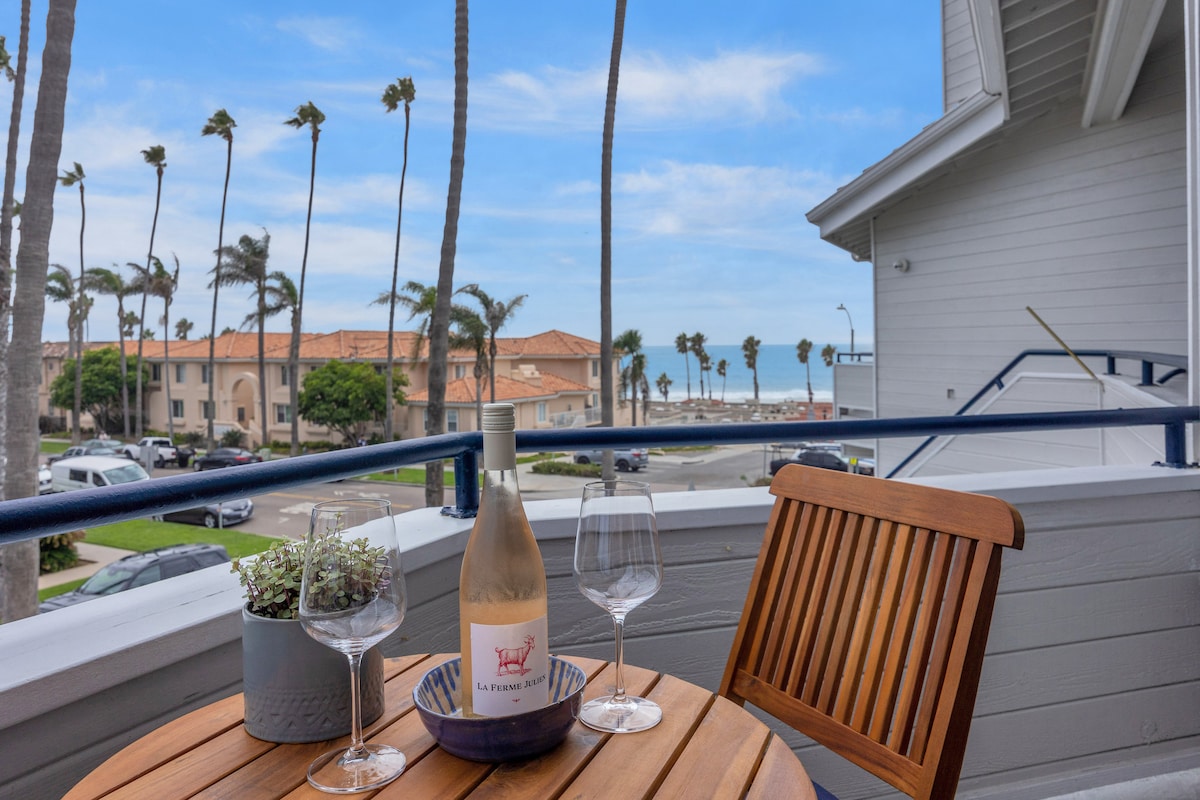 A small outdoor table is set with a bottle of wine and two glasses, surrounded by a scenic view of palm trees and the coastline. Bright blue skies frame the inviting atmosphere, creating a relaxing spot for enjoying refreshments.