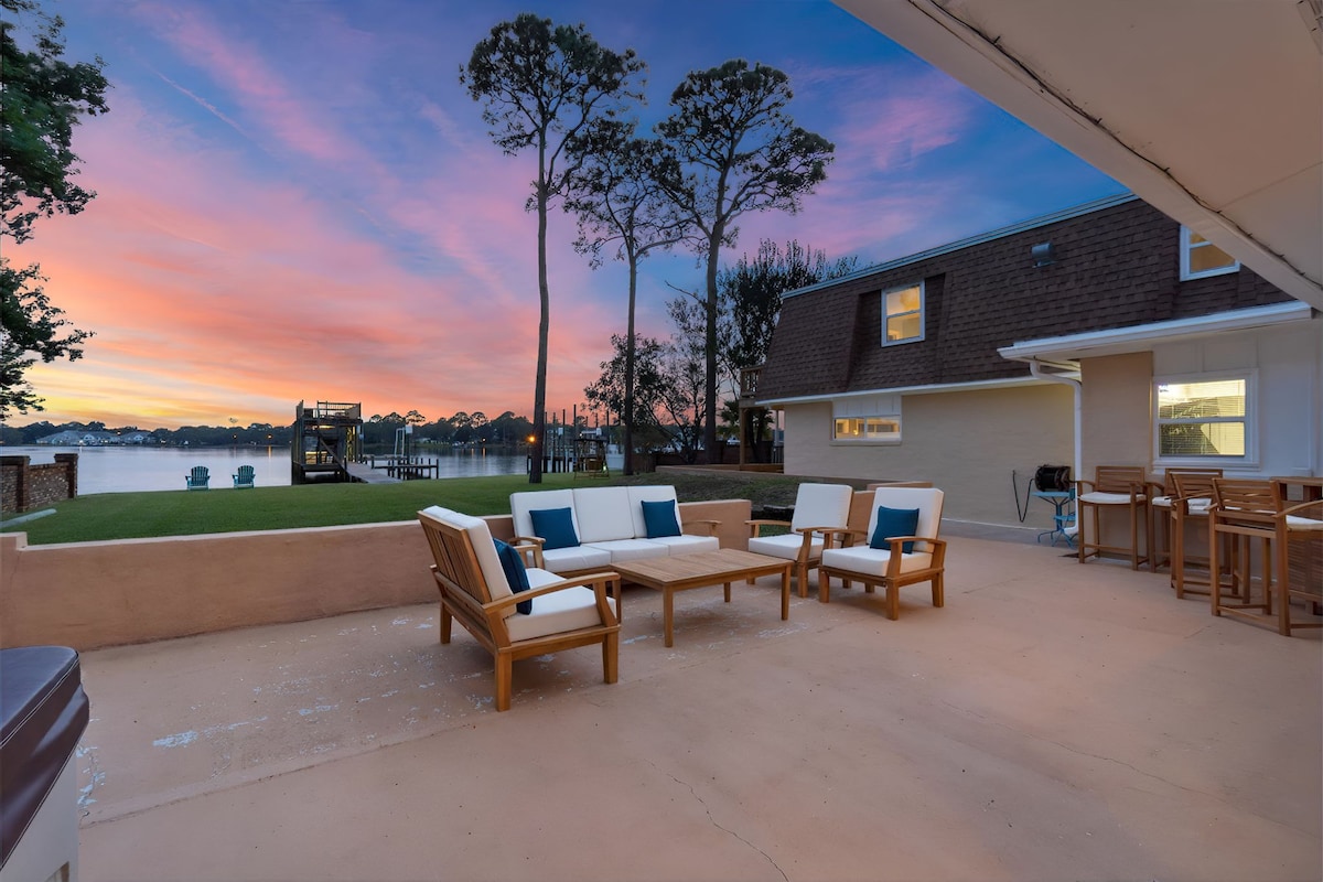An outdoor patio area is furnished with wooden seating and a coffee table, facing the water. The sky is adorned with hues of pink and purple, indicative of sunset, while a dock and trees are visible in the background.