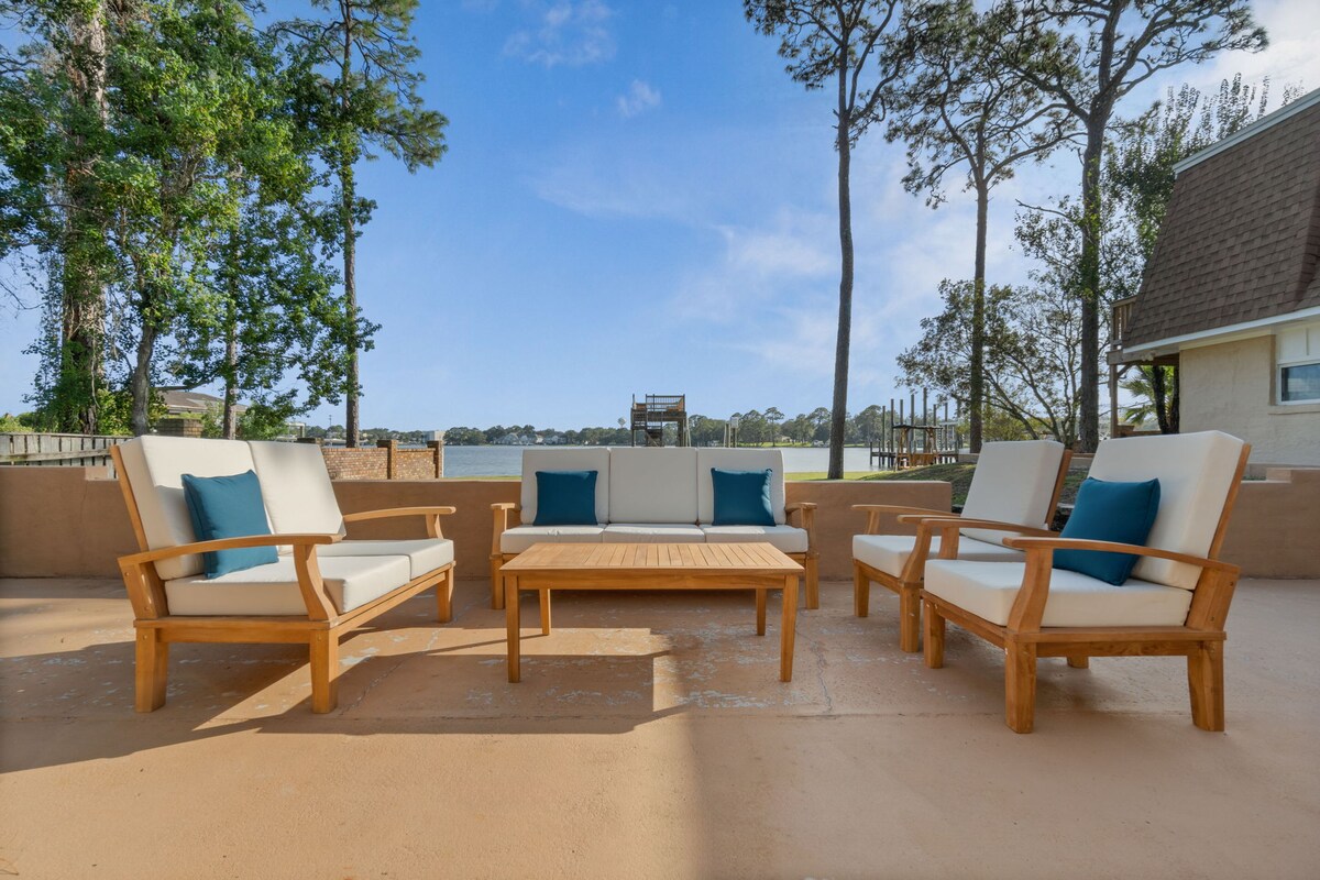 An outdoor patio setting features a wooden coffee table surrounded by four cushioned armchairs, designed for relaxation. A view of calm water is visible in the background, framed by trees and a clear blue sky, creating an inviting space for gatherings.