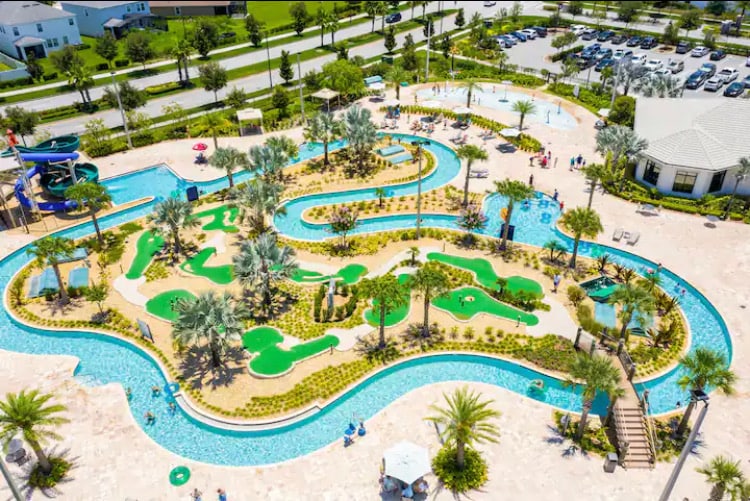Aerial view of a resort-style pool area features winding lazy river and miniature golf course set amidst lush greenery and palm trees. Bright blue water complements the surrounding landscaping, with lounge chairs and umbrellas positioned for relaxation. A community building is visible in the background.